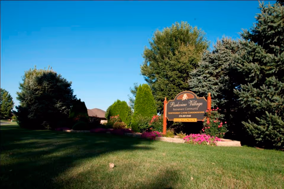 A landscaped outdoor area with green grass, various trees, and flowering plants surrounding a sign that reads 'Parkview Village Retirement Community Independent Living Condominiums' with a phone number and entrance directions.