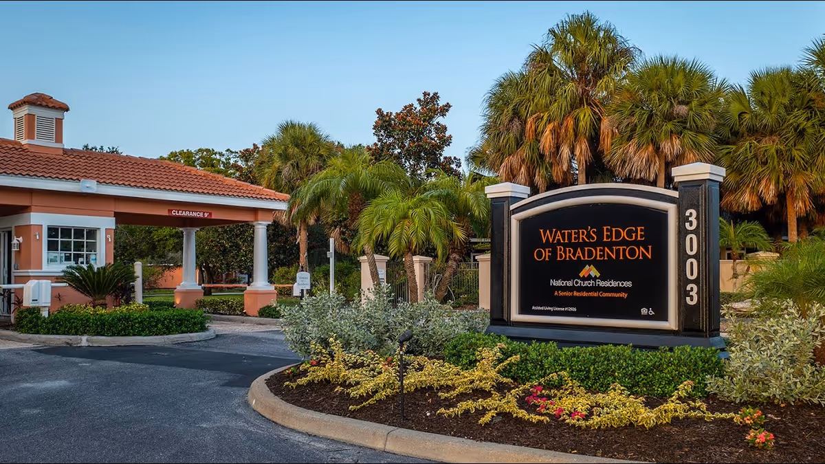 Entrance of Water's Edge of Bradenton with sign, palm trees, and curved driveway.