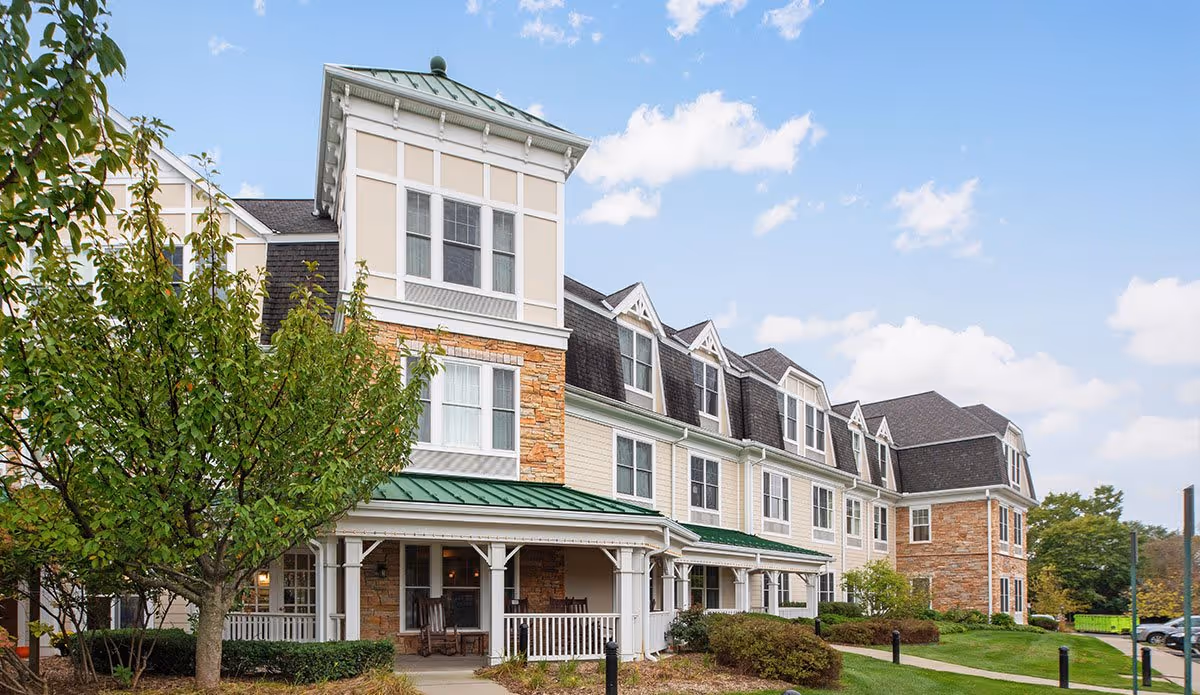 Exterior view of a multi-story senior living facility building with a green metal roof, white and beige siding, stone accents, and multiple windows. There is a covered porch with rocking chairs, a tree, shrubs, and a well-maintained lawn under a partly cloudy sky.
