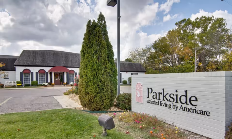 Front entrance and lawn of Parkside assisted living with a brick sign, evergreen tree, and building façade.