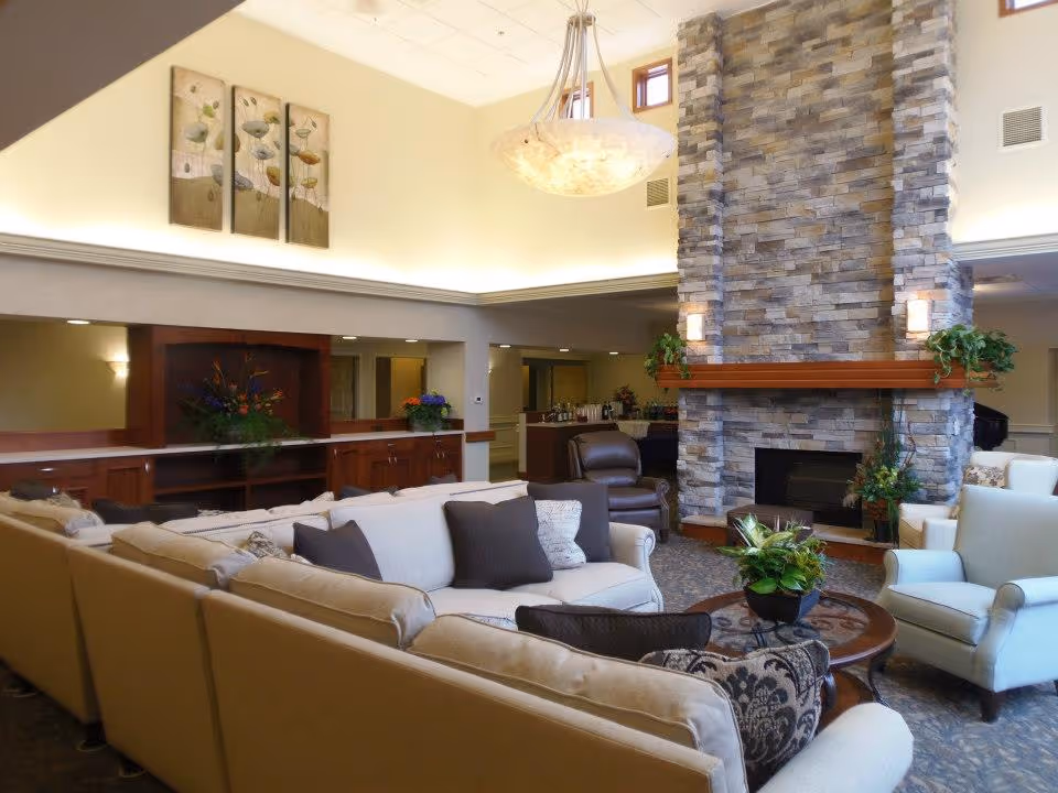 A spacious and well-lit living room area with beige sofas and armchairs arranged around a round wooden coffee table with a plant centerpiece. The room features a tall stone fireplace with a wooden mantel decorated with green plants. Above the seating area hangs a large, modern chandelier. The walls are light-colored, and there is a decorative triptych painting on one wall. In the background, there are wooden cabinets and a counter with flowers and other decorative items.