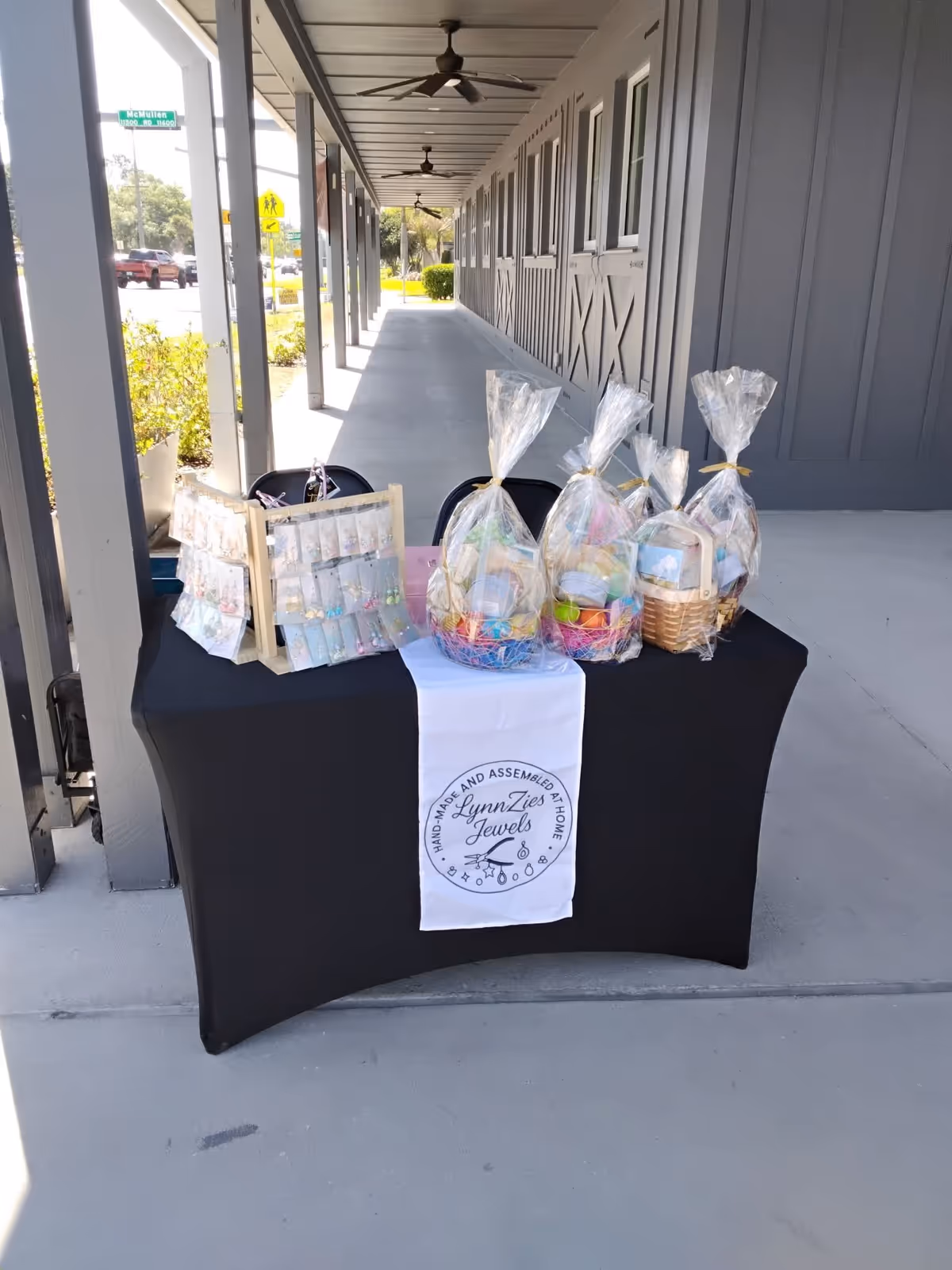 Table with gift baskets wrapped in clear plastic on a black tablecloth along an outdoor walkway.
