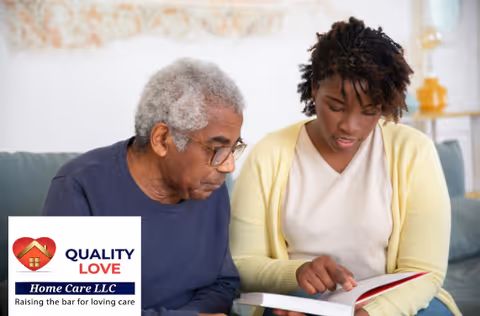 An elderly man and a caregiver look at a book together on a living room couch.
