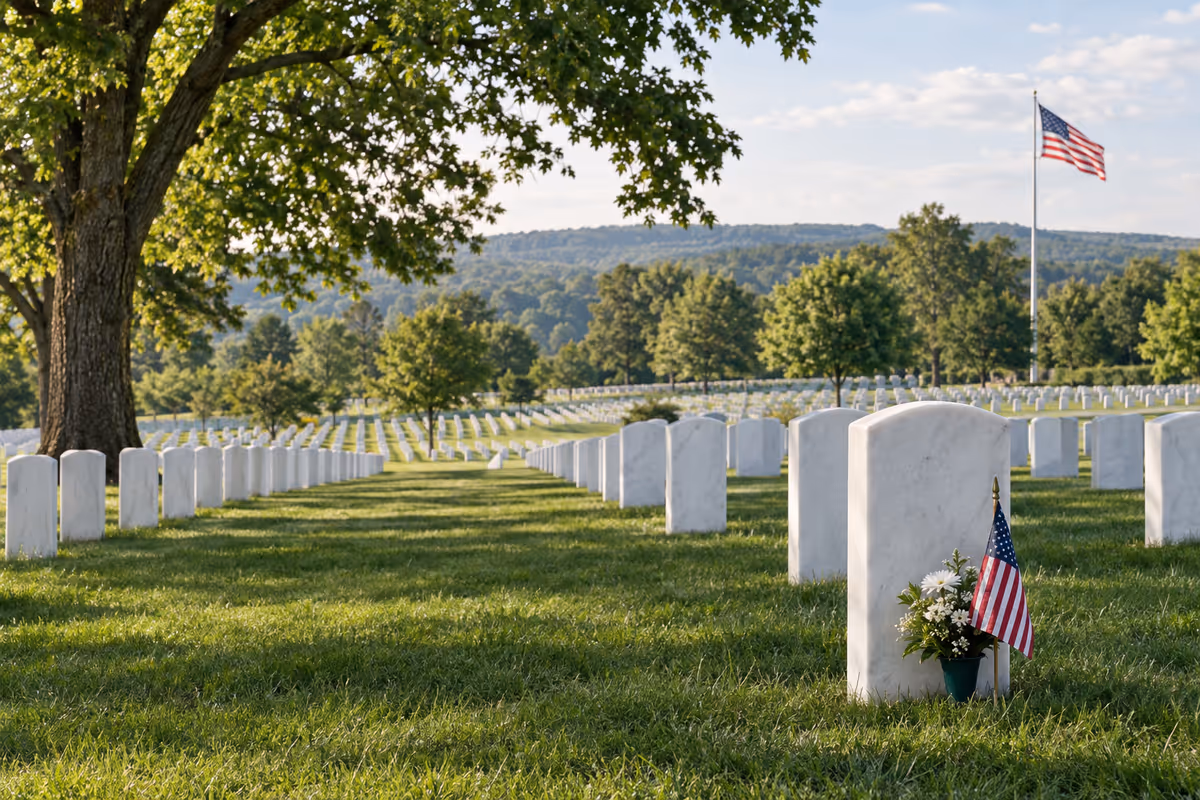 Rows of white headstones in a national cemetery, with small American flags and a flag in the distance