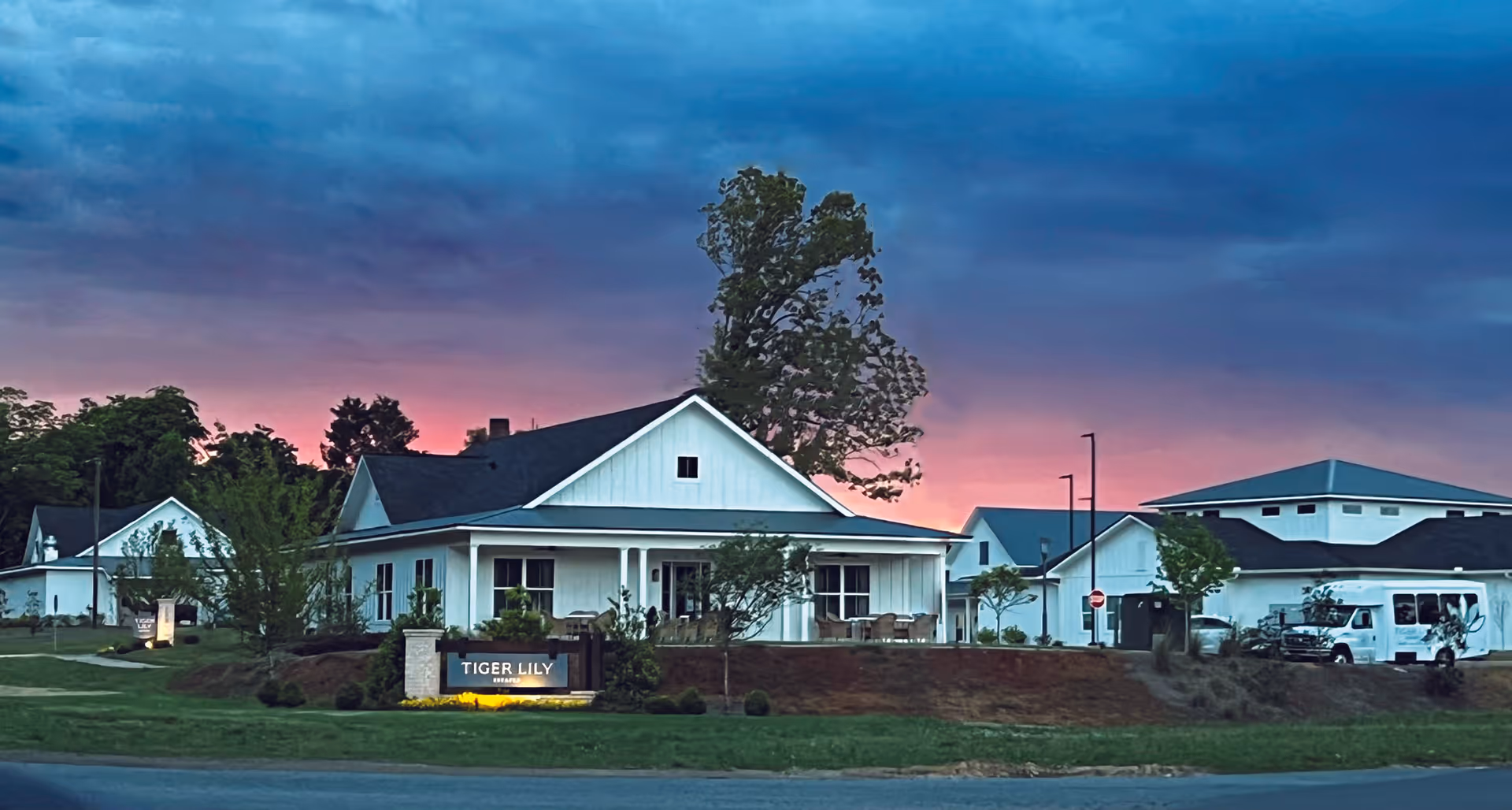 Exterior view of Tiger Lily Estates Assisted Living & Memory Care facility at dusk with a partly cloudy sky showing shades of blue and pink. The building is white with a dark roof, surrounded by landscaped greenery and trees. A sign in front reads 'Tiger Lily Estates'.