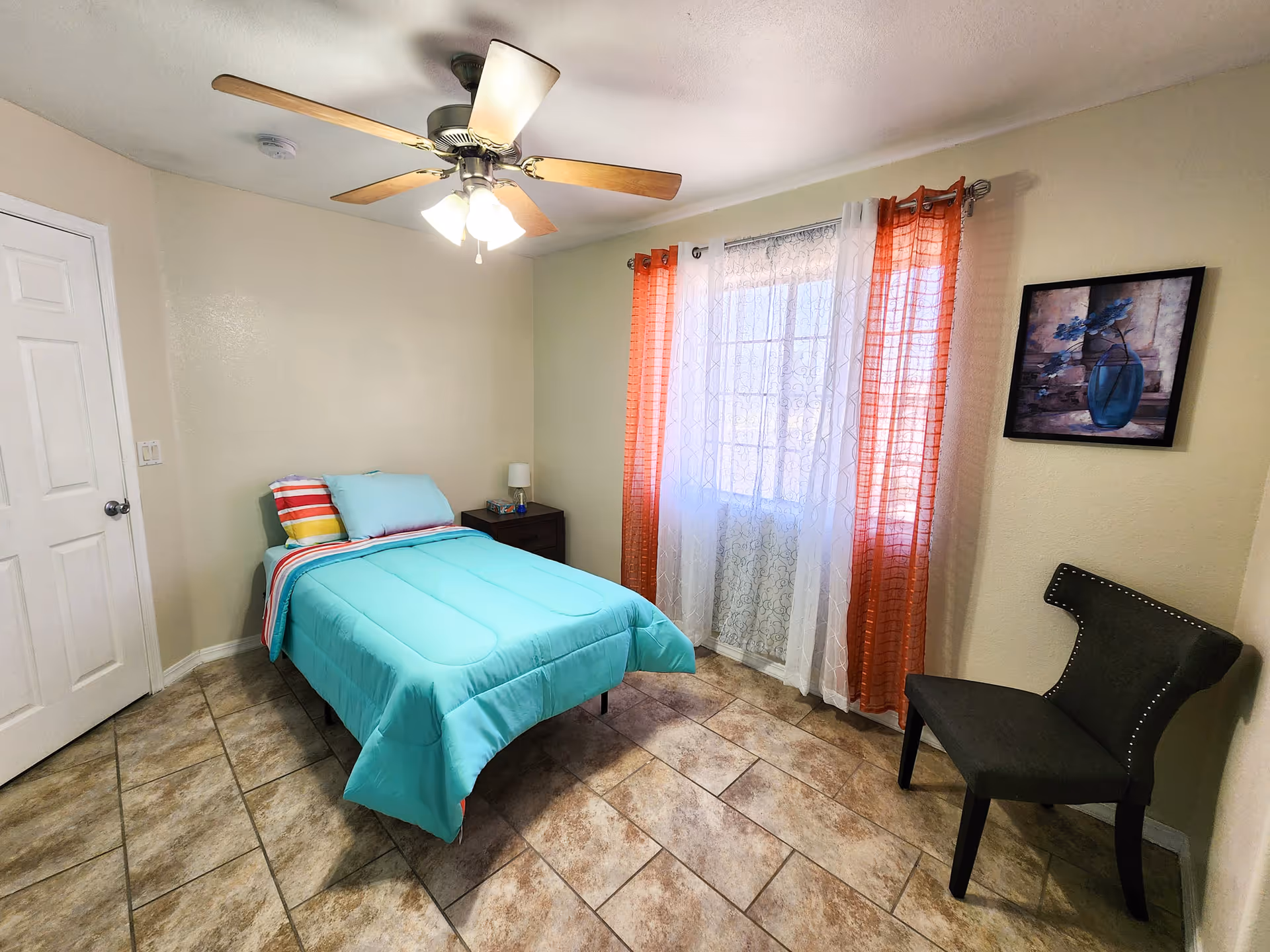 Small bedroom with turquoise bedspread, ceiling fan, window with sheer orange curtains, a nightstand, and chair.