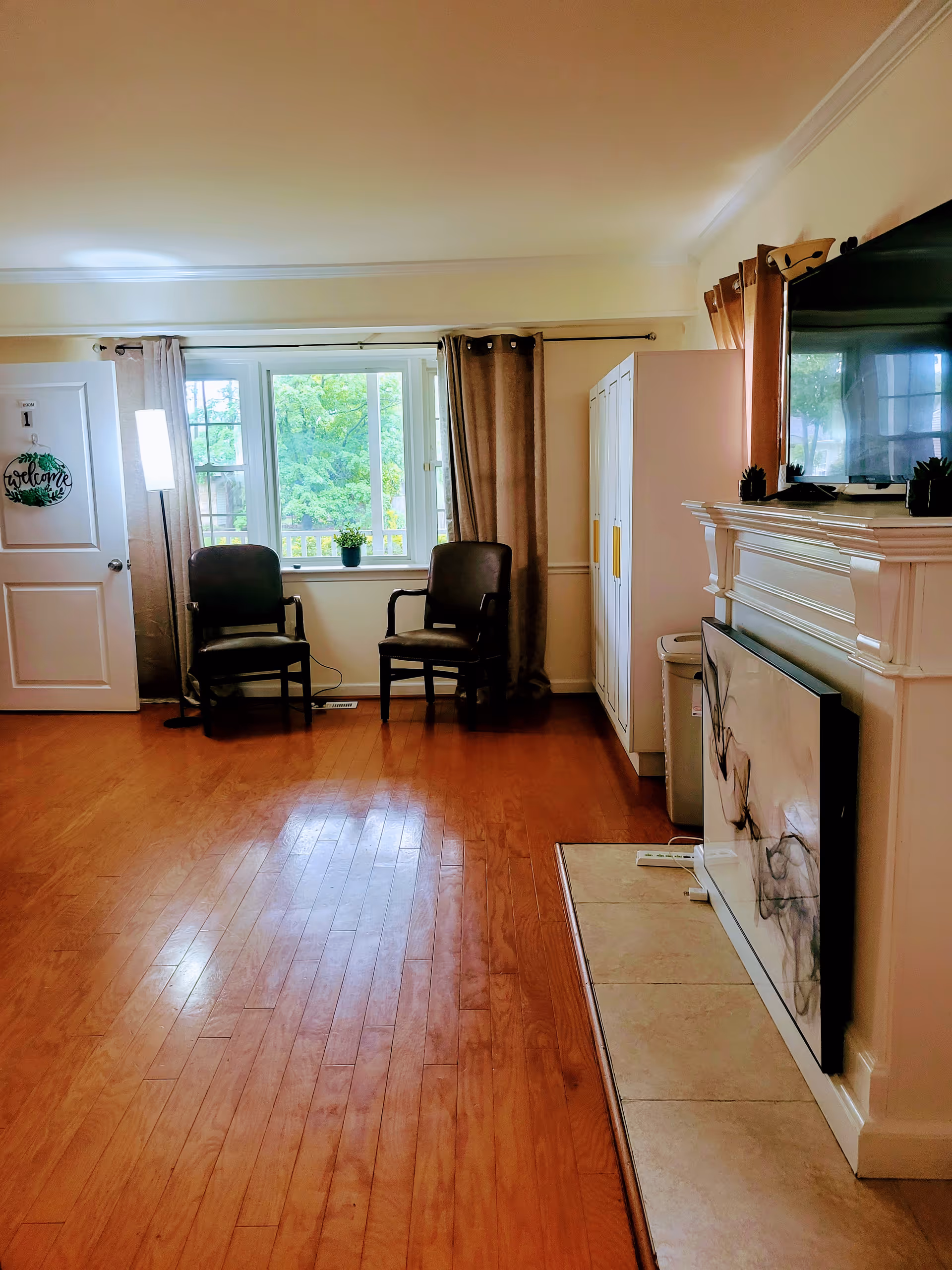 Cozy living room with two chairs, window, door with welcome sign, and TV on a mantel.