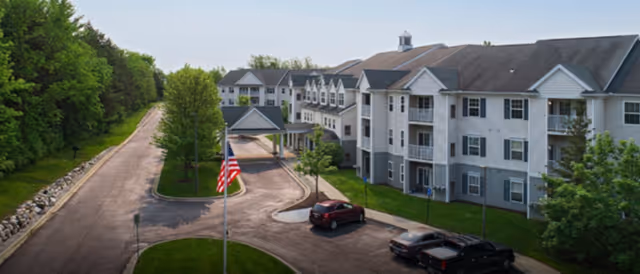 Multi-story senior living building with driveway, flag, parked cars, and landscaped grounds.