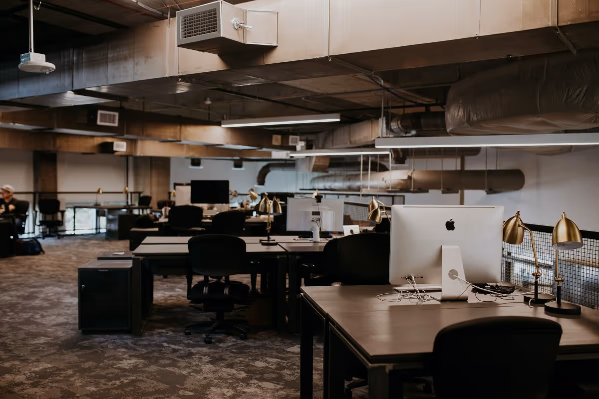 Industrial open-office space with desks, chairs, iMacs, and exposed ceiling ducts.