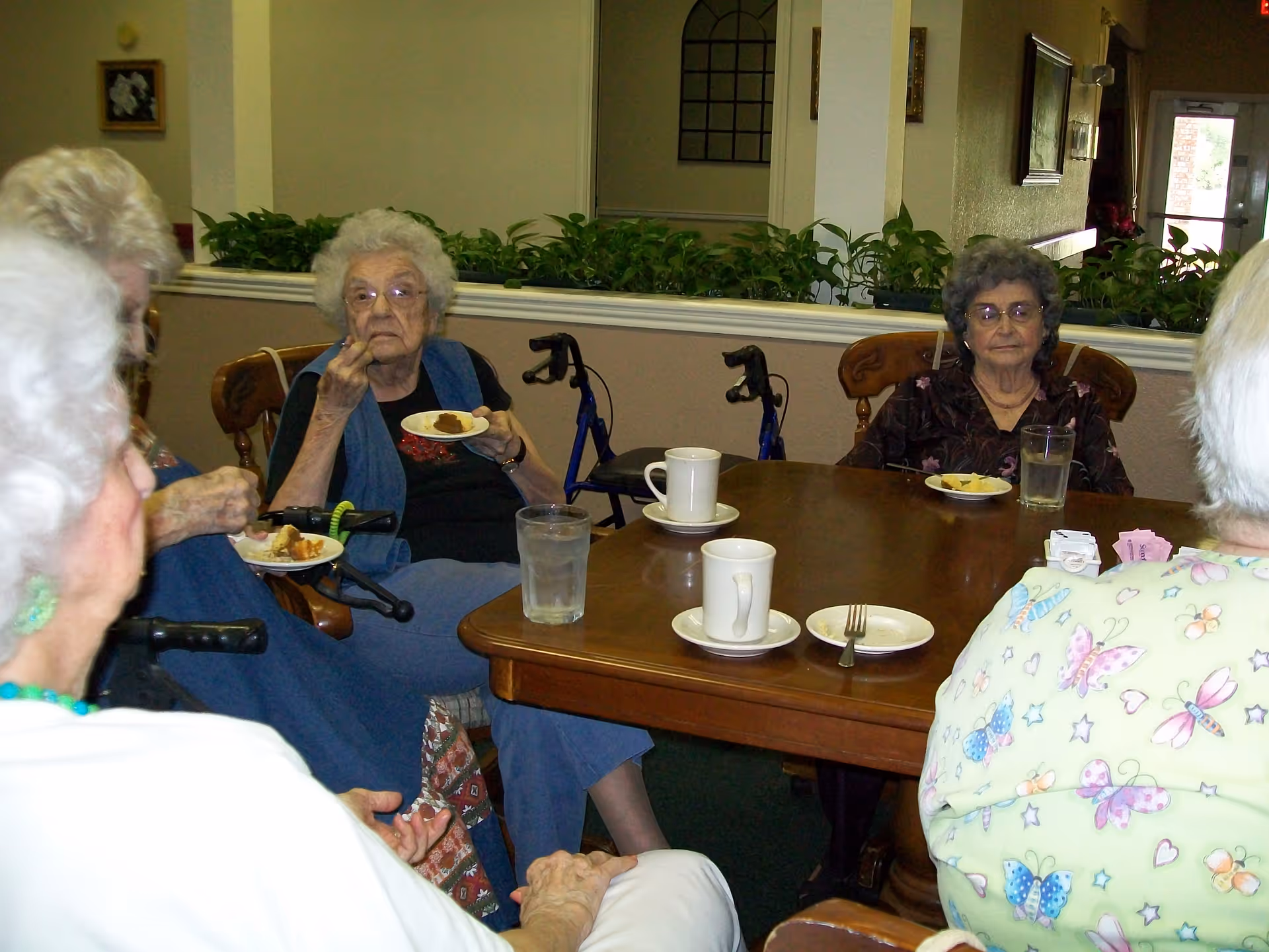 A group of elderly women sitting around a wooden table in a communal dining area, eating and drinking. The room has plants on a ledge behind them and a door with a window in the background.