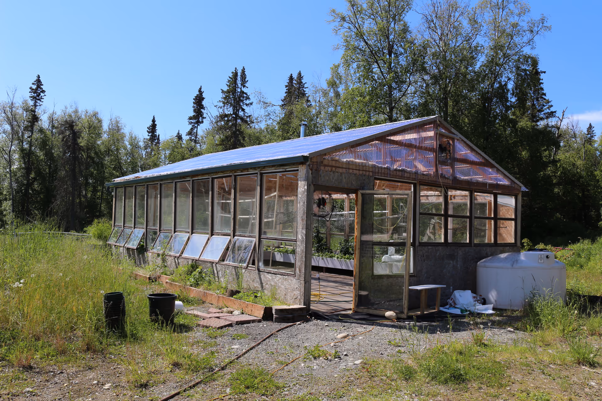 A weathered greenhouse with glass panels sits in a grassy rural area beside a water tank.