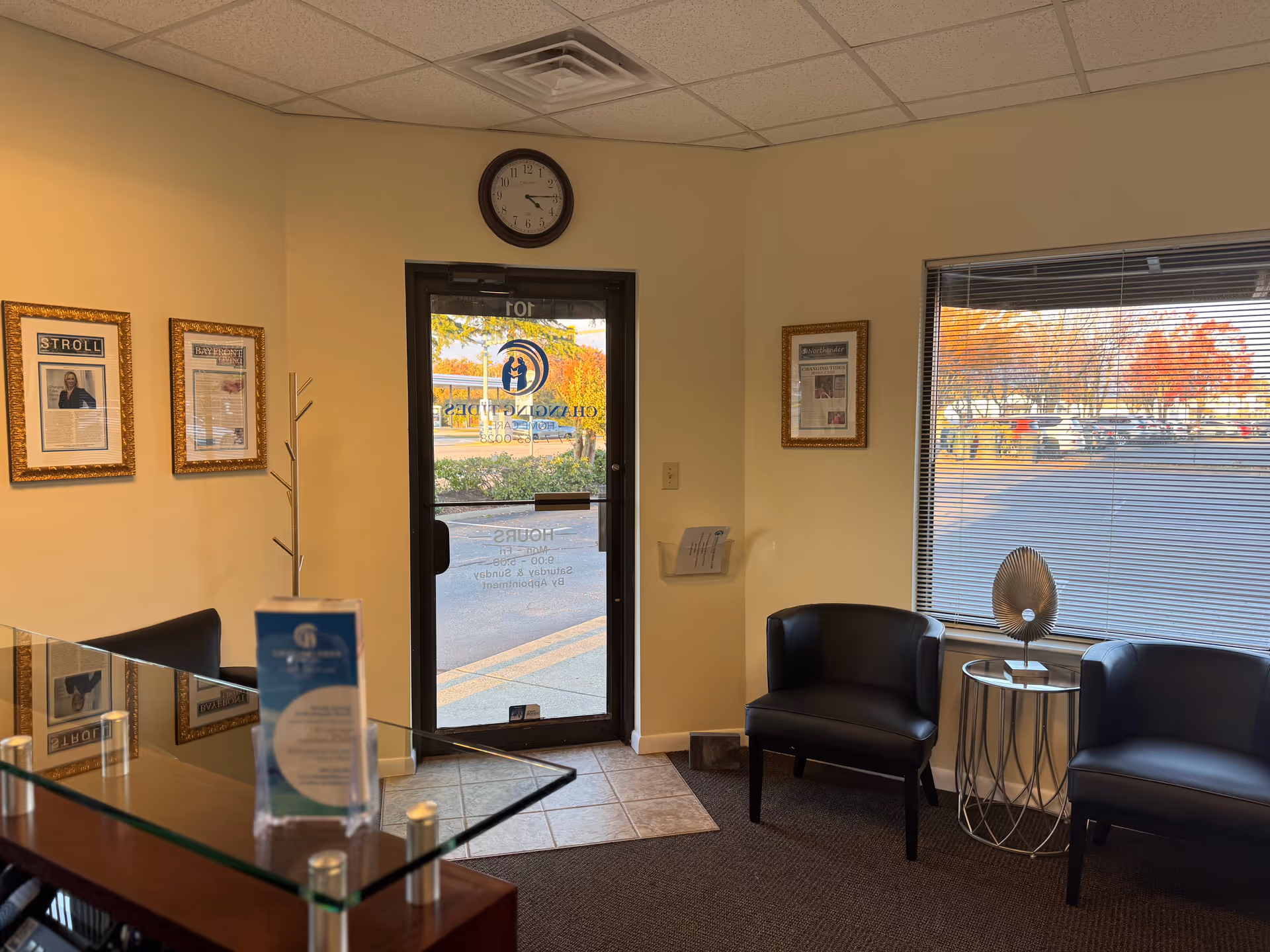 Interior lobby of Changing Tides Home Care with glass desk, clock, framed notices, and black chairs.