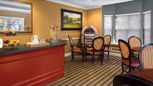 Bright senior living dining area with a red counter, round tables, cushioned chairs, and a jukebox.