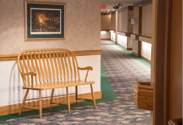 A hallway in an assisted living facility with patterned carpet and green borders. A wooden bench is placed against the wall on the left side, beneath a framed painting. The hallway is softly lit with wall sconces and has an exit sign visible in the distance.