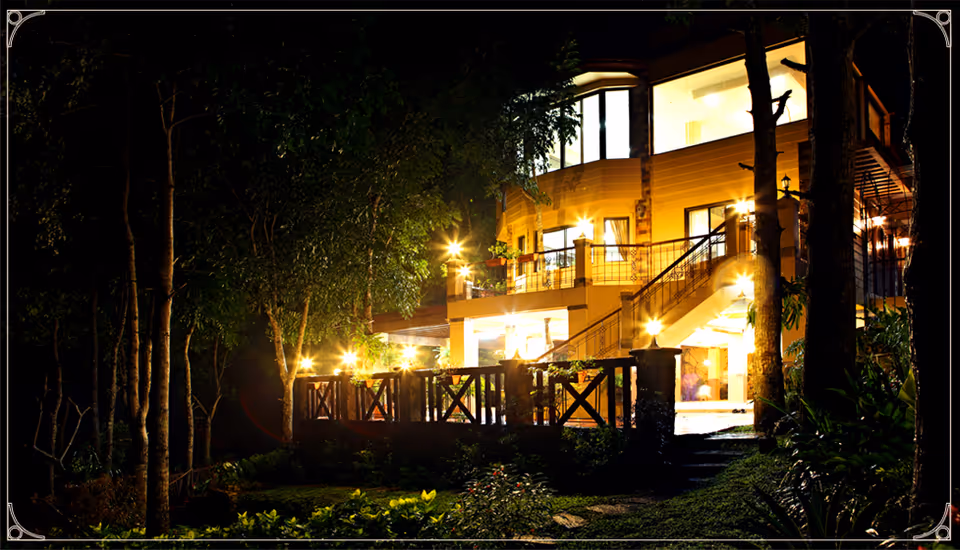 Nighttime exterior of a multi-level building with illuminated balconies, stairs, and a wooden deck surrounded by trees.