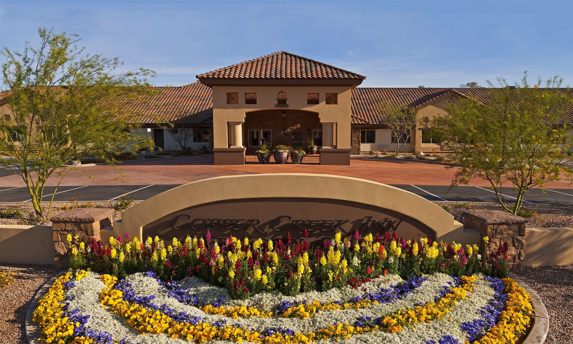 Front entrance of Copper Creek Inn with a curved sign and colorful flowerbeds in front of a tile-roofed building.