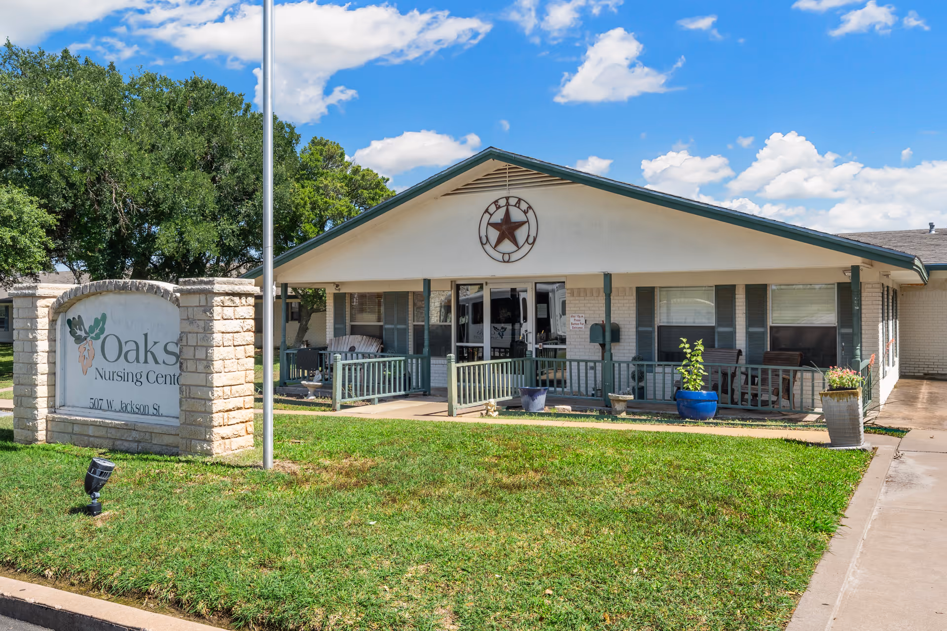 Front exterior view of Oaks Nursing Center building with a porch, green railing, potted plants, and a sign displaying the facility's name and address. The sky is blue with some clouds and there are trees in the background.