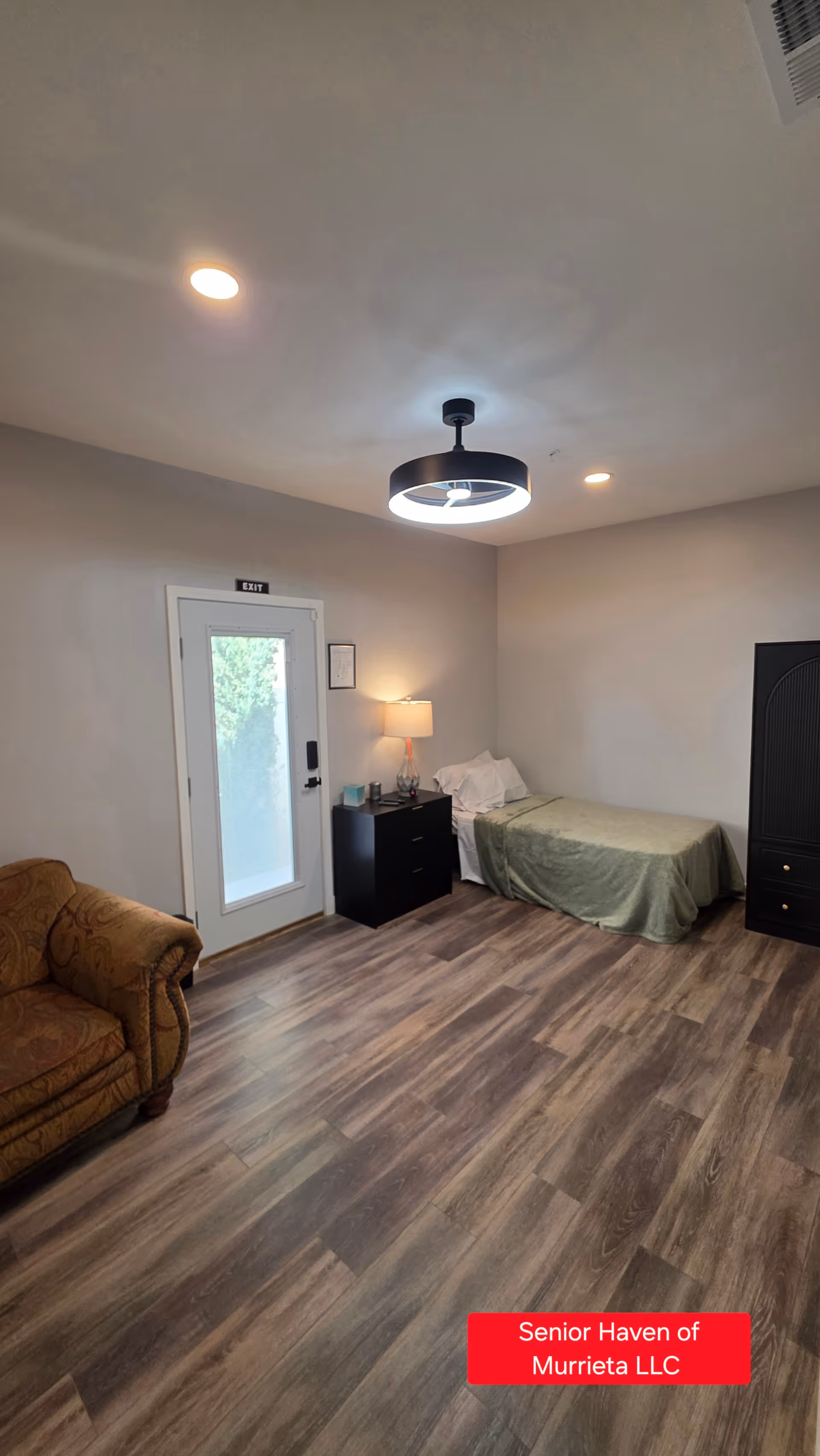 Spacious bedroom with bed, couch, nightstand, and exit door in a senior living facility.