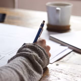Close-up of a hand writing on documents at a wooden table with a mug.