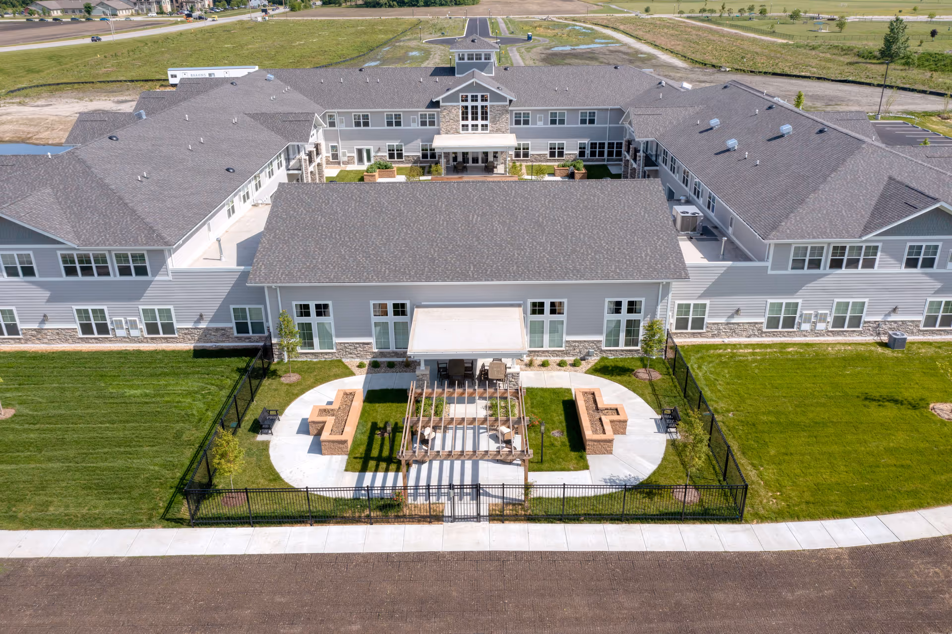 Aerial view of a two-story U-shaped senior living building with a fenced courtyard, pergola seating area, and surrounding lawn.