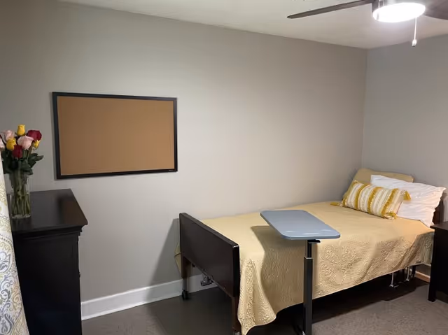 Small bedroom with a single bed, dresser, flowers, and a corkboard on gray wall.