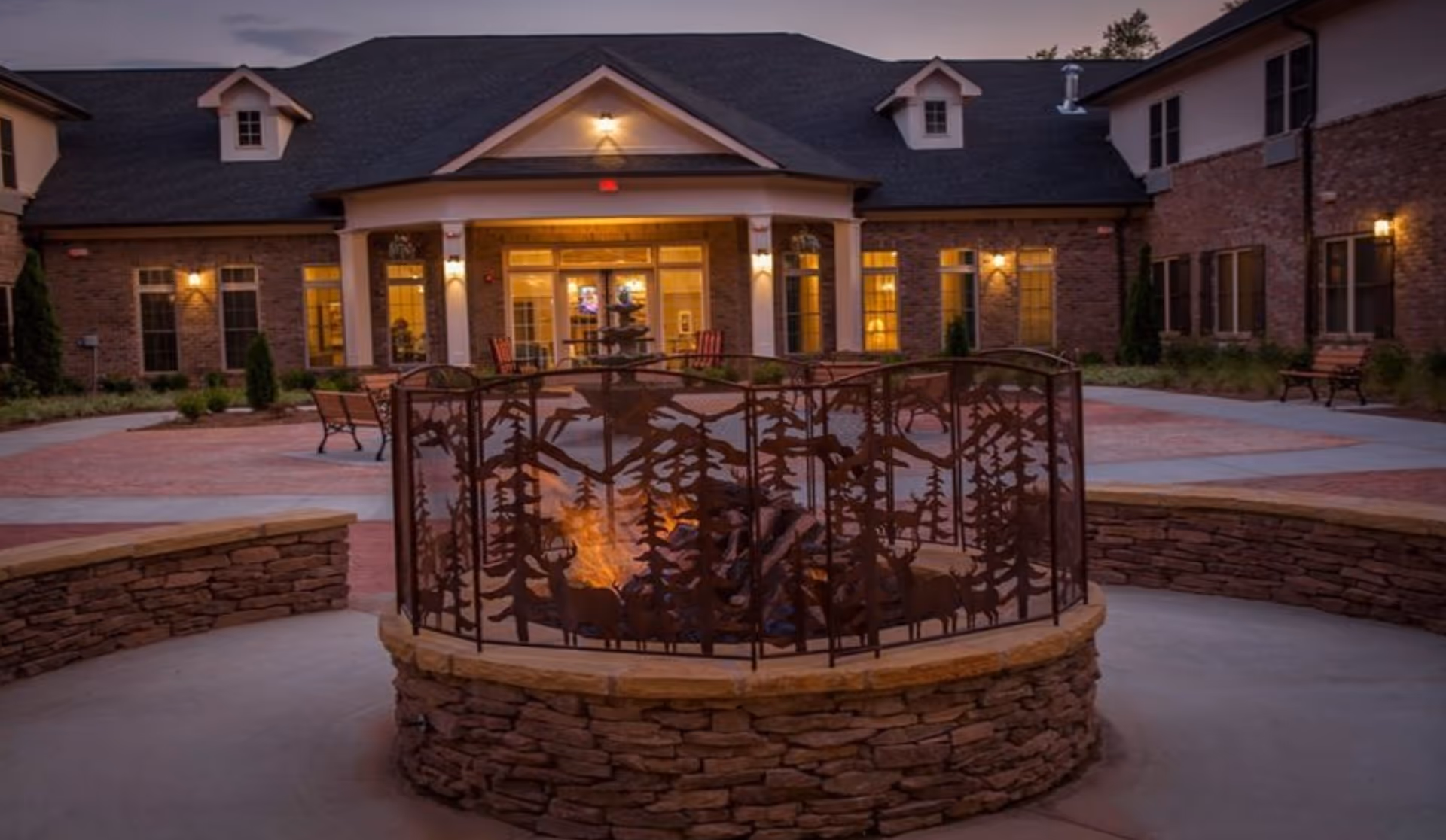 Exterior courtyard at dusk with a stone fire pit surrounded by a decorative metal screen and the lit entrance of the building in the background.