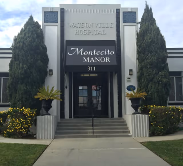 Entrance of Montecito Manor building with steps leading to double doors, a black awning sign, and tall trees on either side.