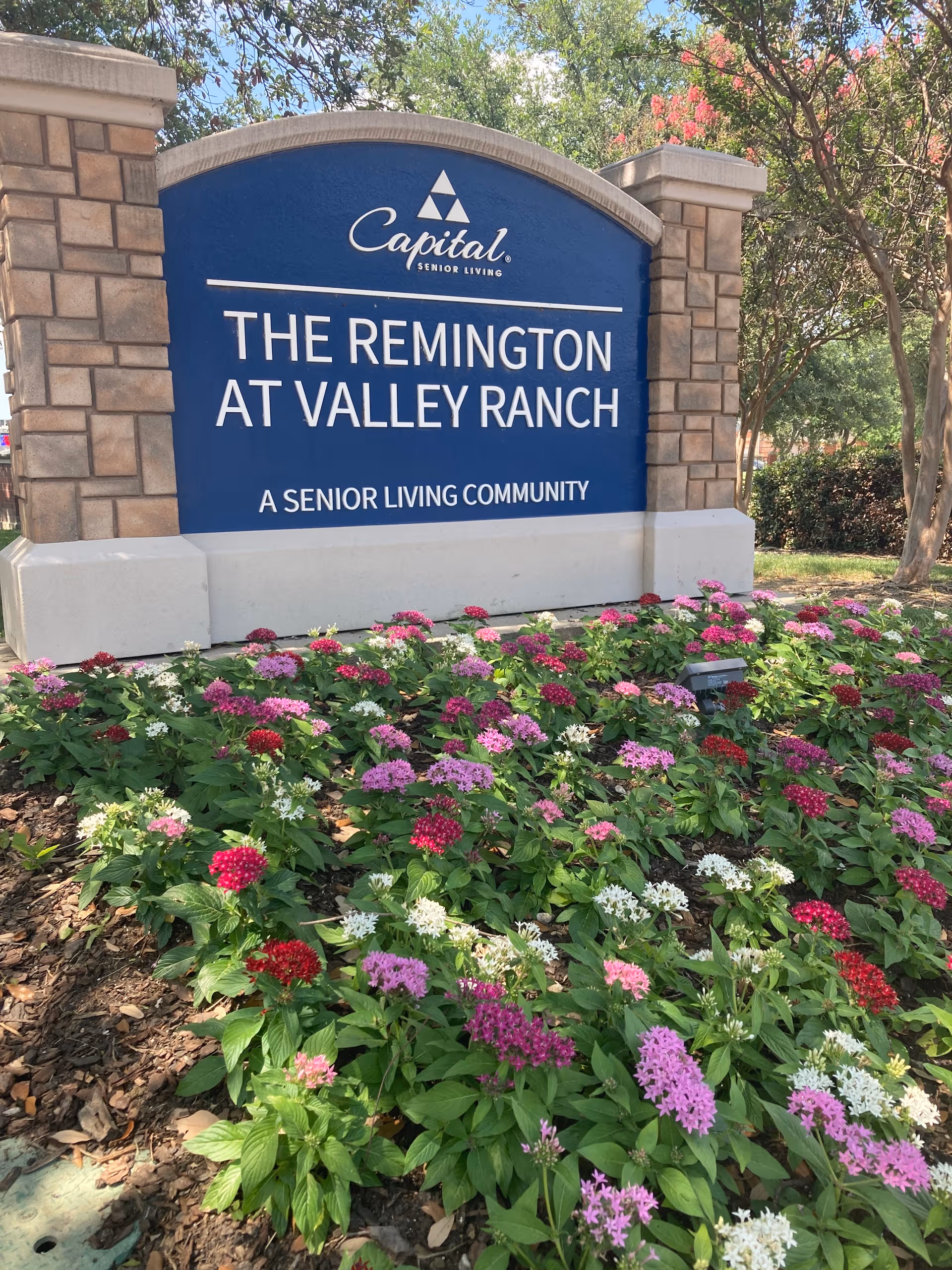 Blue sign reads The Remington at Valley Ranch, Capital Senior Living, with colorful flowers in front.