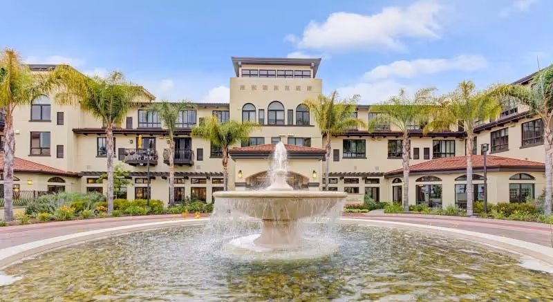 Front exterior view of Merrill Gardens at Campbell, featuring a multi-story building with arched windows, palm trees, and a large circular fountain in the foreground under a partly cloudy sky.