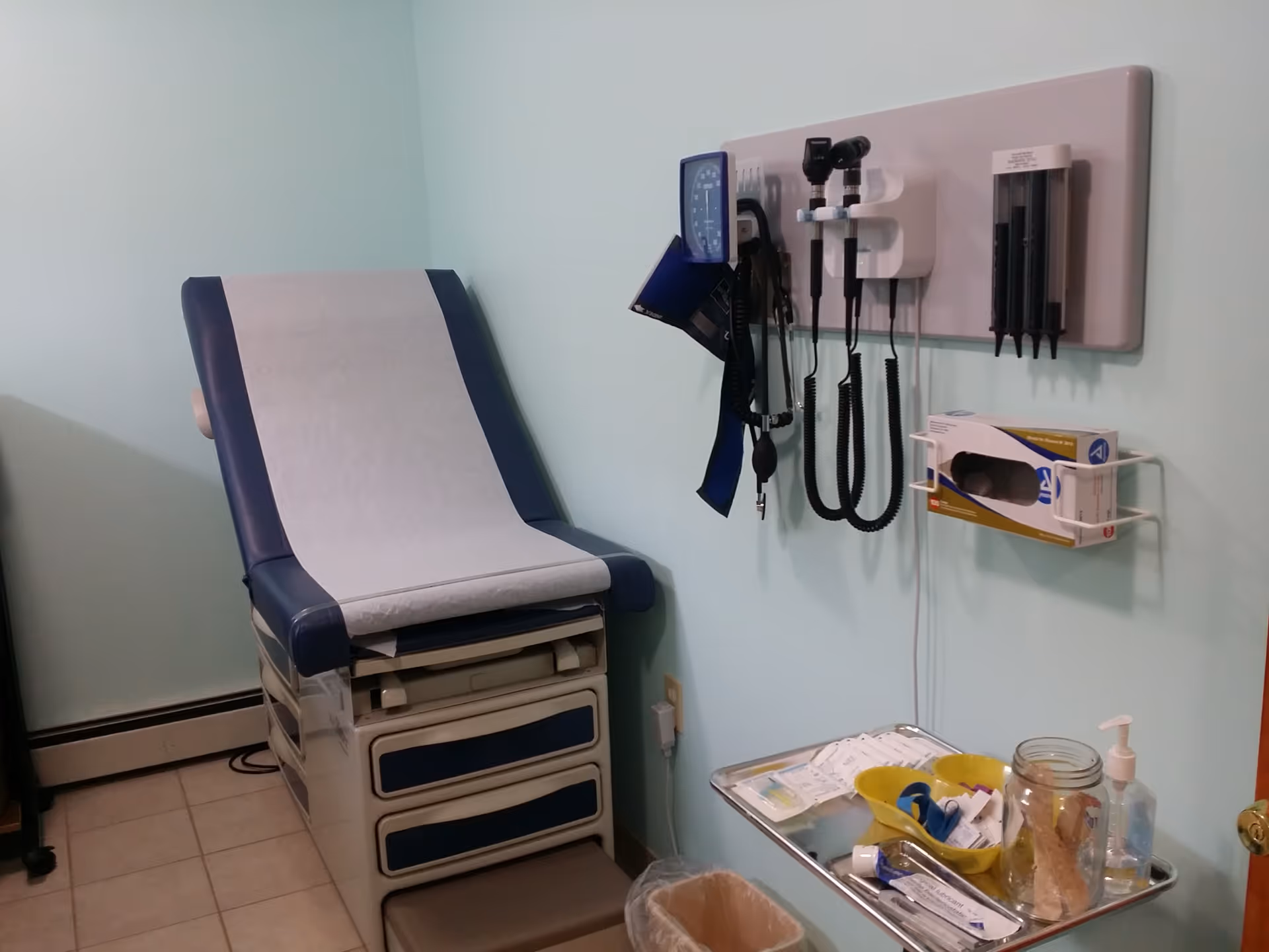 Medical exam room with blue adjustable chair, wall-mounted equipment, and a tray of supplies.