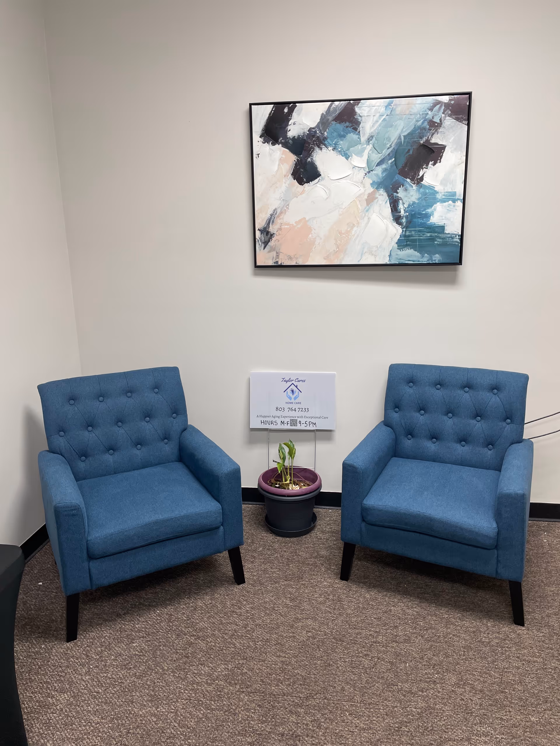 Two blue tufted chairs, a potted plant, and abstract wall art in a small lounge.