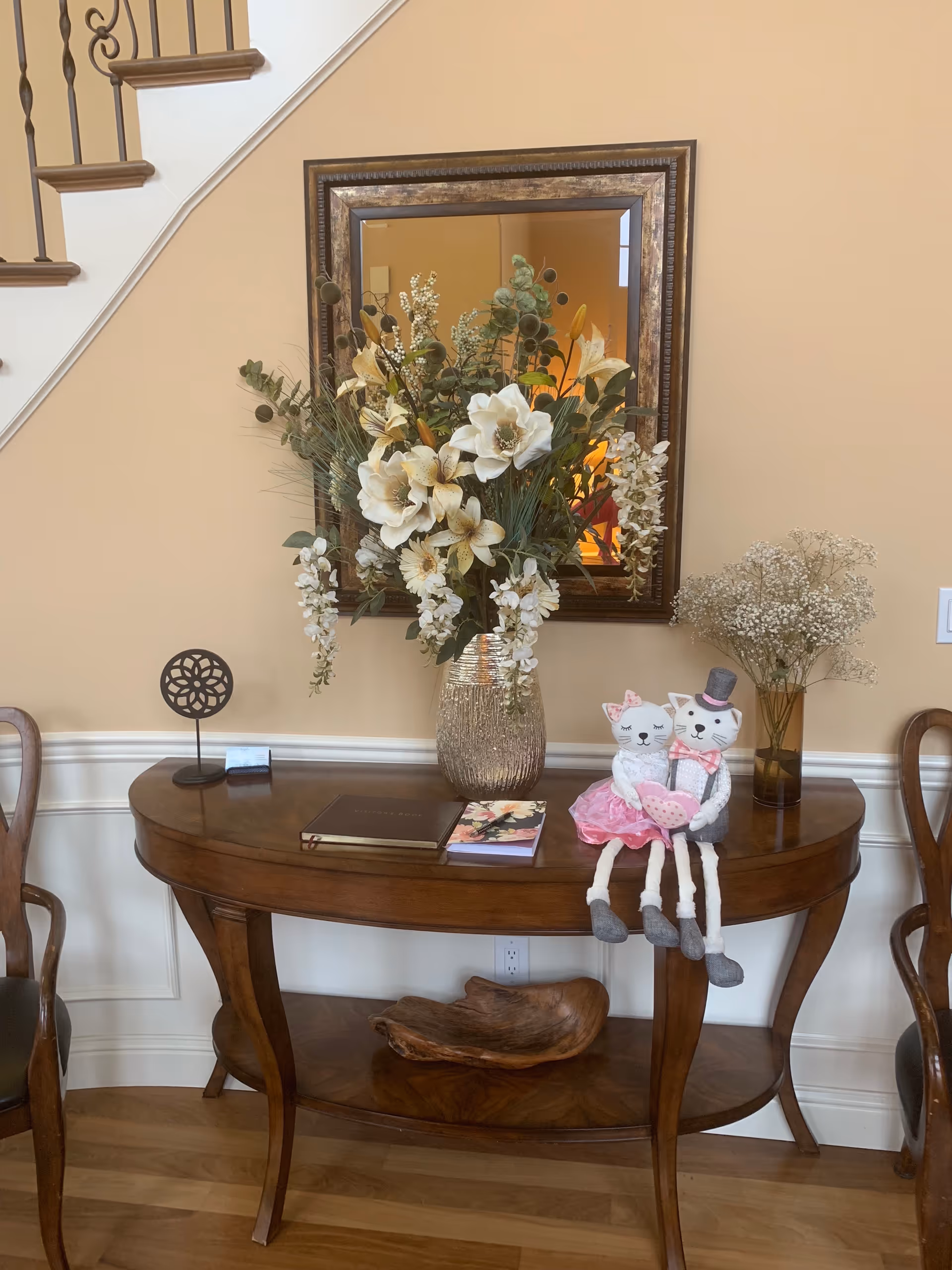 Wooden console table with floral arrangement, mirror, and two stuffed cat dolls in a beige foyer.