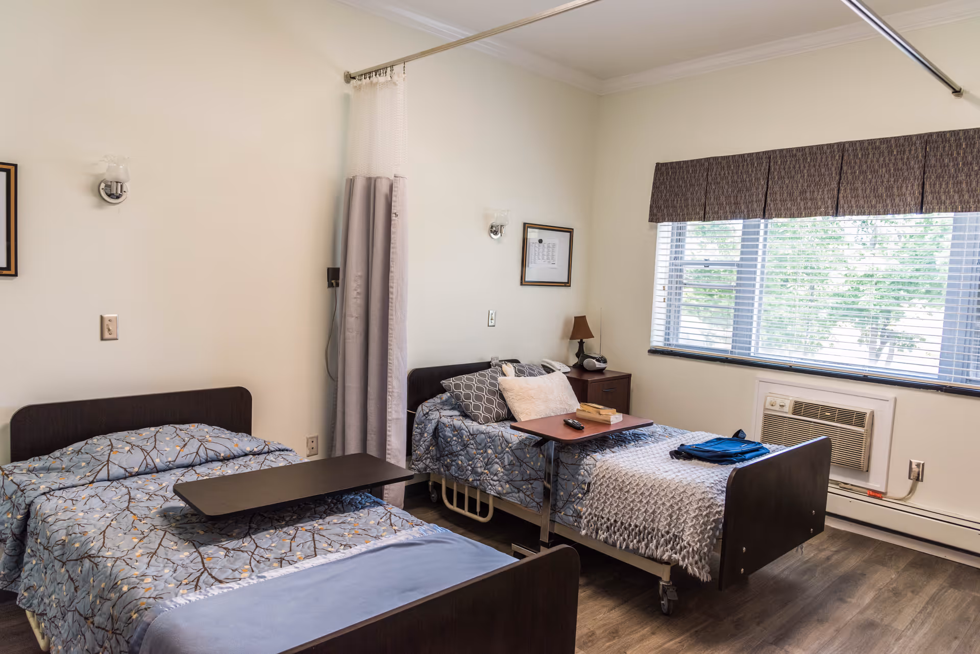 A room with two single beds, each with patterned bedding and a small overbed table. One bed has a pillow and a blanket, and the other has a pillow and a folded blanket. There is a window with blinds and a valance, an air conditioning unit below the window, a small bedside table with a lamp and a phone, and a privacy curtain between the beds. The floor is wood, and the walls are light-colored.