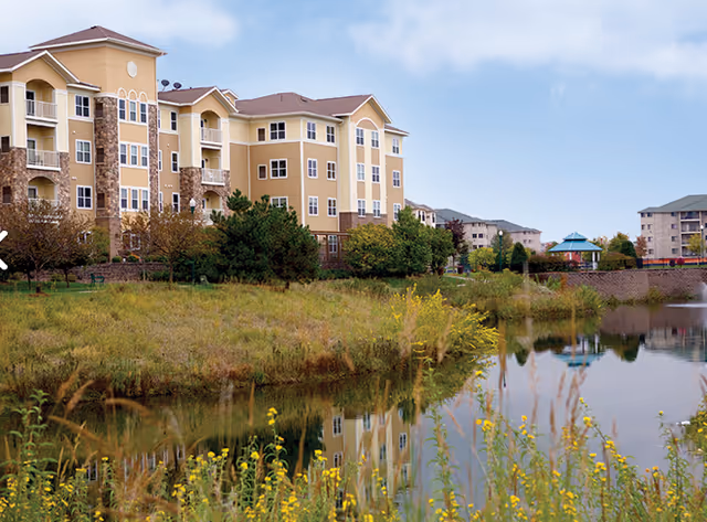 Multi-story beige senior living building beside a reflective pond with trees and yellow wildflowers.