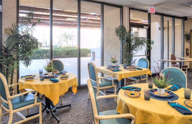 Dining area with round tables covered in yellow tablecloths and place settings near large glass windows and an exit door.