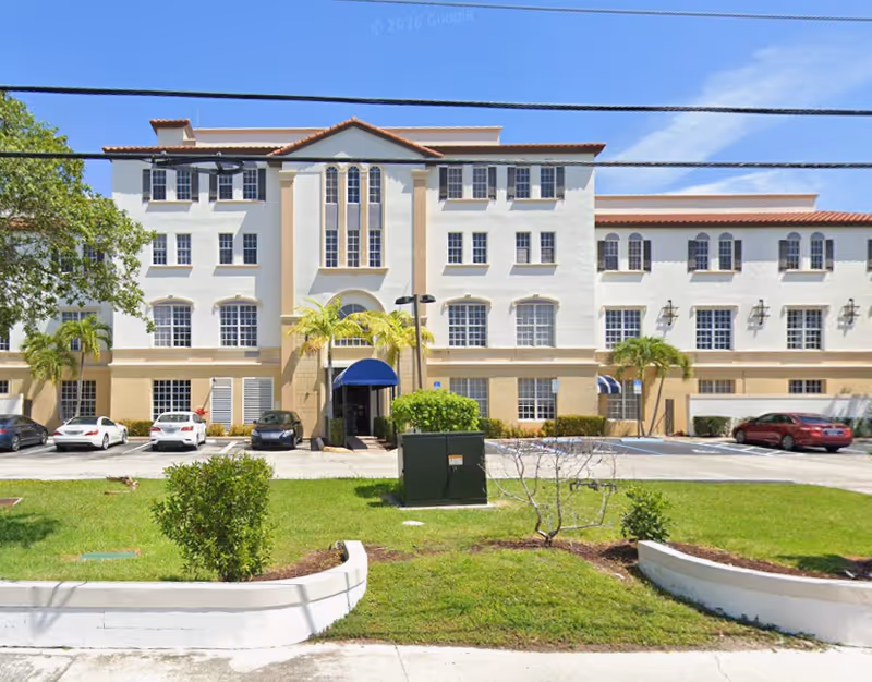 White multi-story senior living building with beige accents, blue entrance awning, and palm trees.