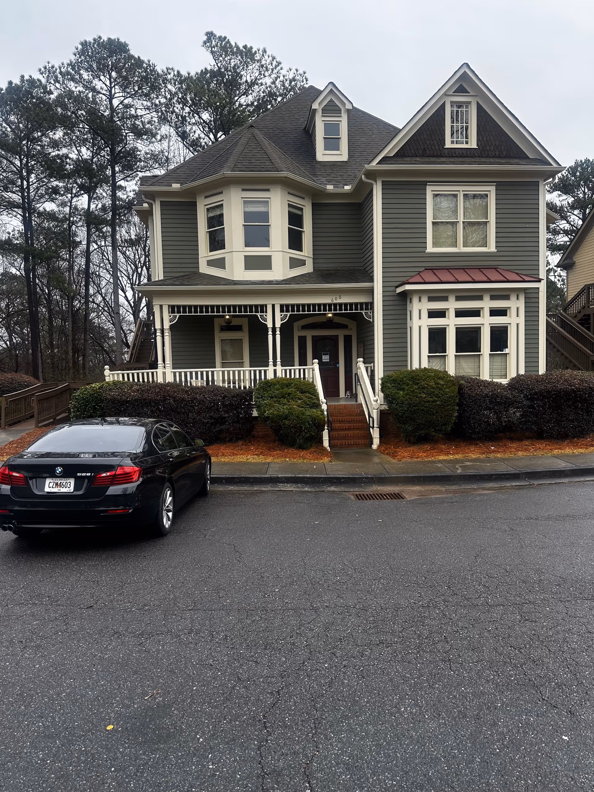 Two-story gray house-style building with front porch, landscaping, and a parked car nearby.