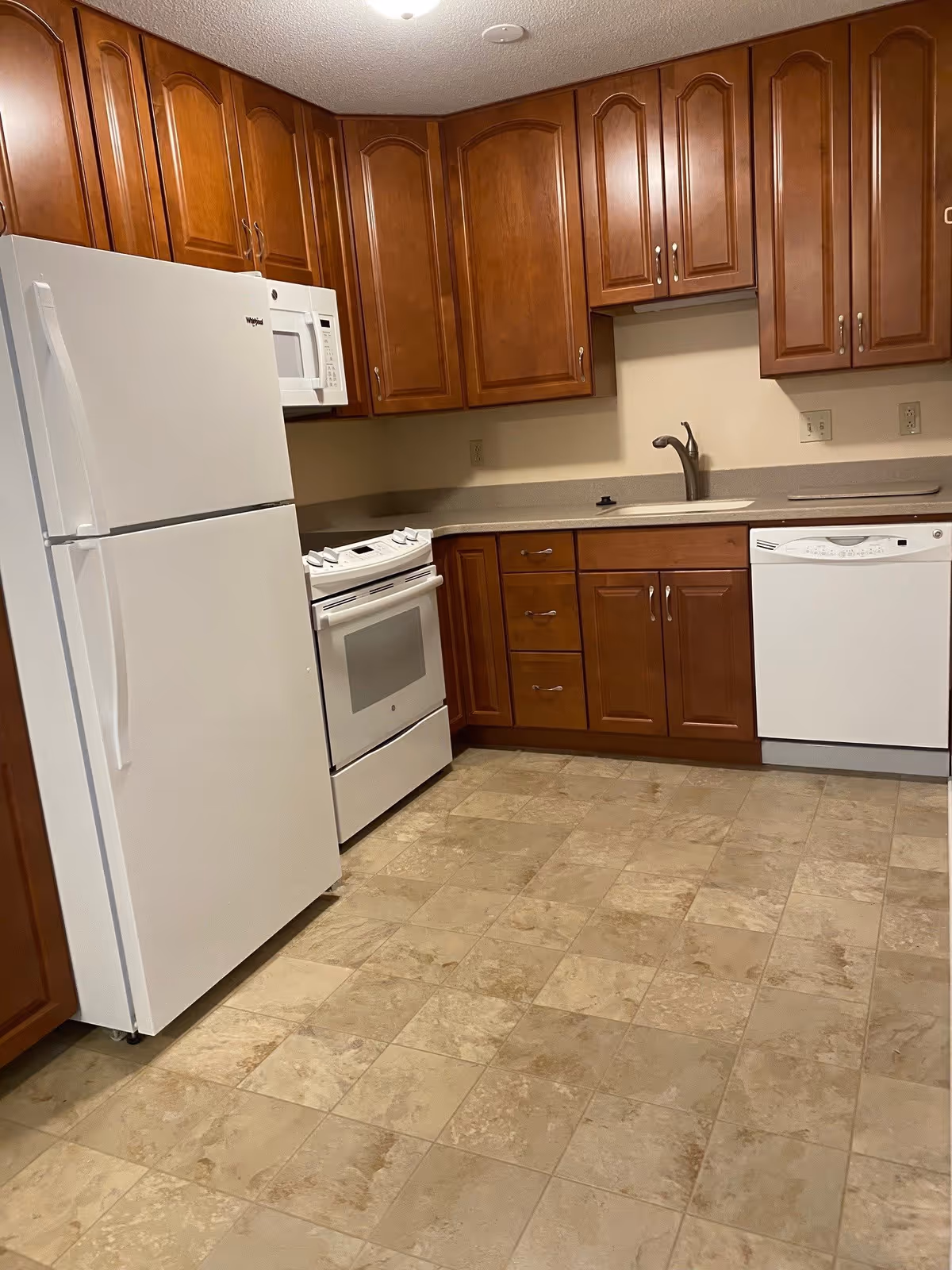 A kitchen with wooden cabinets, a white refrigerator, a white microwave above a white stove, a white dishwasher, a sink with a faucet, and beige tiled flooring.