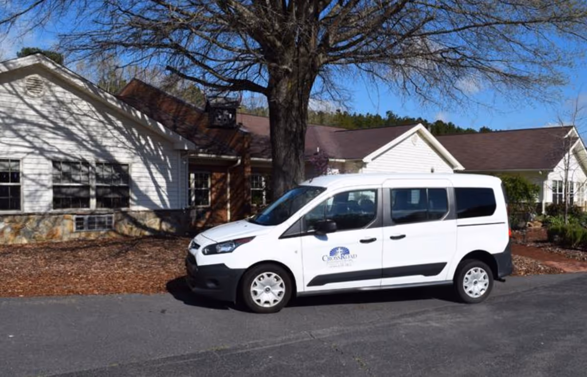 A white Cross Road–branded van parked in front of a single-story retirement community building beneath a large tree.
