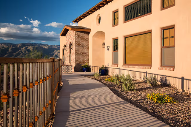 A paved walkway leading to the entrance of a beige stucco building with brick accents, surrounded by a metal railing and landscaped with small plants and flowers. Mountains and a partly cloudy sky are visible in the background.