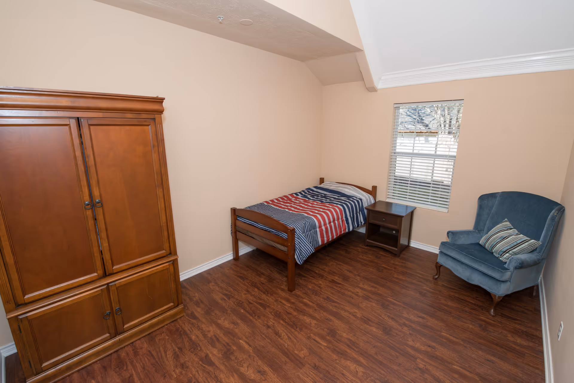A small bedroom with wooden flooring, a single bed with a striped red, white, and blue blanket, a wooden nightstand, a blue upholstered armchair with a striped pillow, and a large wooden wardrobe. There is a window with white blinds letting in natural light.