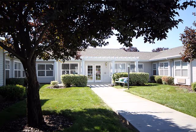 Single-story senior living building front with a paved walkway leading through a grassy courtyard to glass double doors, flanked by shrubs and a bench.