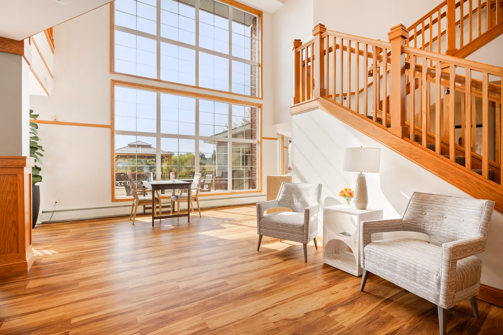 Bright and spacious interior area with large floor-to-ceiling windows letting in natural light. Two cushioned armchairs are placed on either side of a small white side table with a lamp and a small flower arrangement. Wooden staircase with railing is visible on the right side. Wooden flooring and light-colored walls create a warm and inviting atmosphere. A small table with chairs is positioned near the windows, and an outdoor gazebo can be seen through the windows.