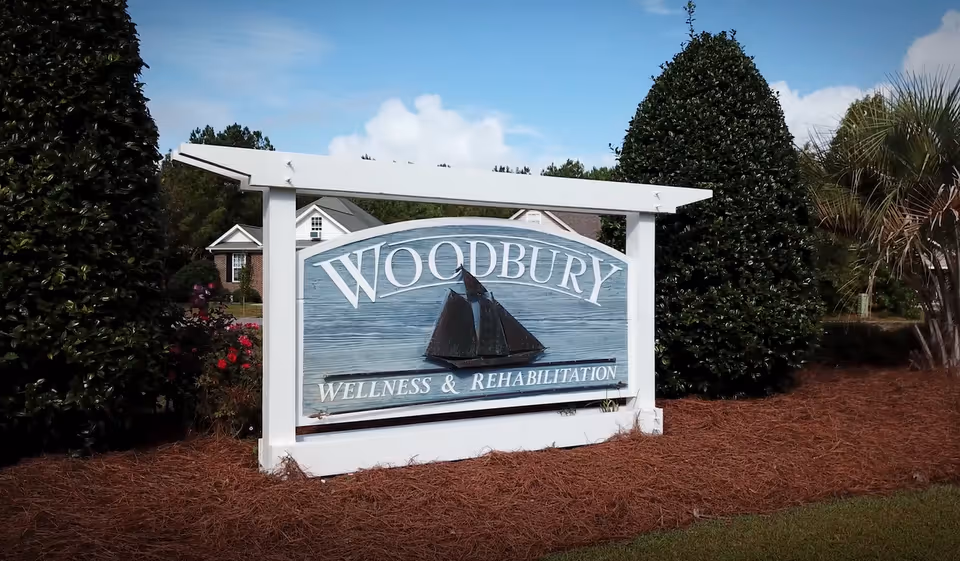 Woodbury Wellness & Rehabilitation sign outdoors, framed by shrubs and reddish mulch.