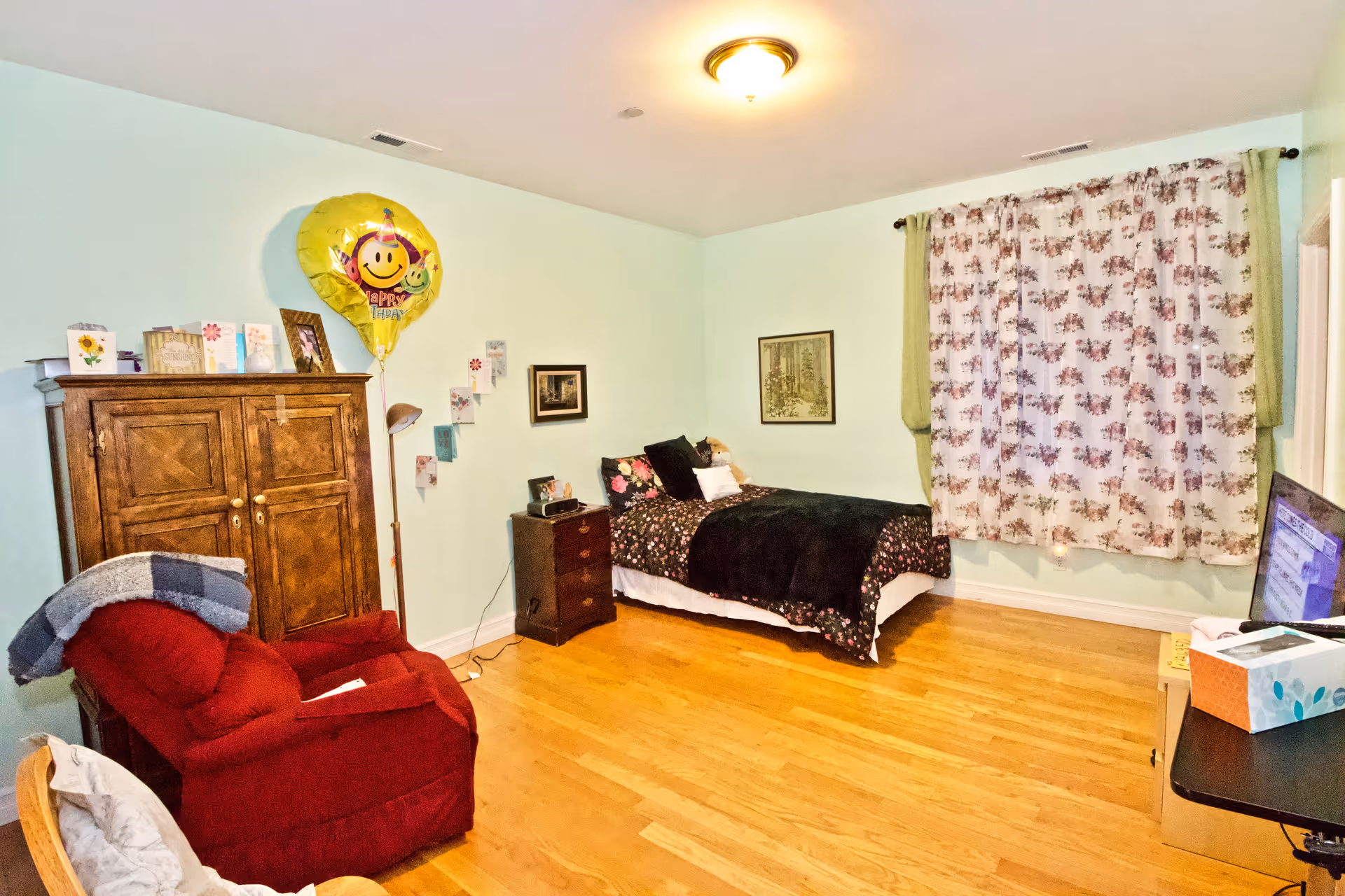 A cozy bedroom in a senior living facility with bed, dresser, chair, and TV.