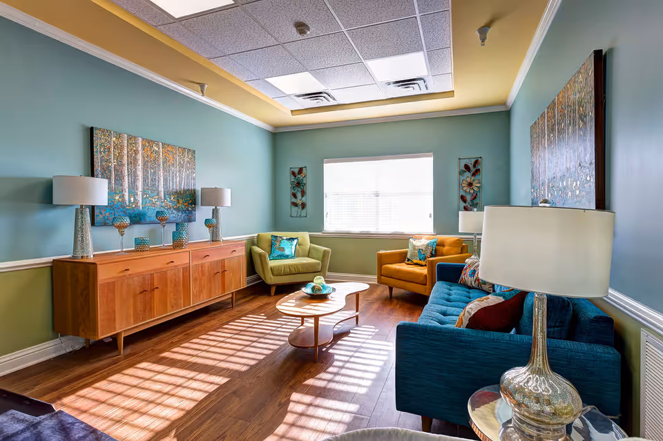Bright lounge area with teal walls, colorful couches, and wooden sideboard in a senior living facility.