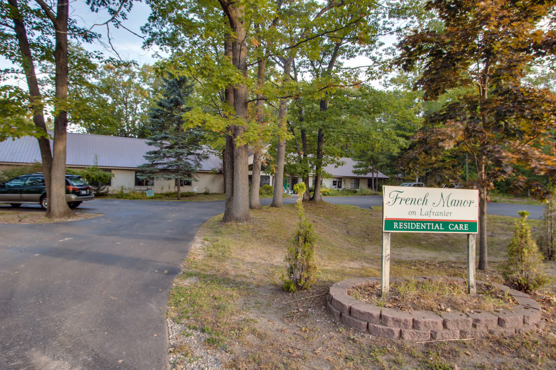 Sign for 'French Manor' residential care in front of a single-story building surrounded by trees and a driveway.