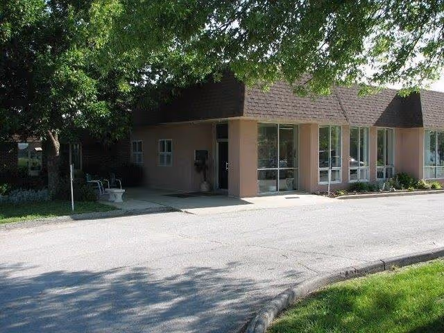 Exterior view of a single-story building with large windows and a brown shingled roof, partially shaded by a large tree. There is a paved driveway or road in front and some green grass on the side.