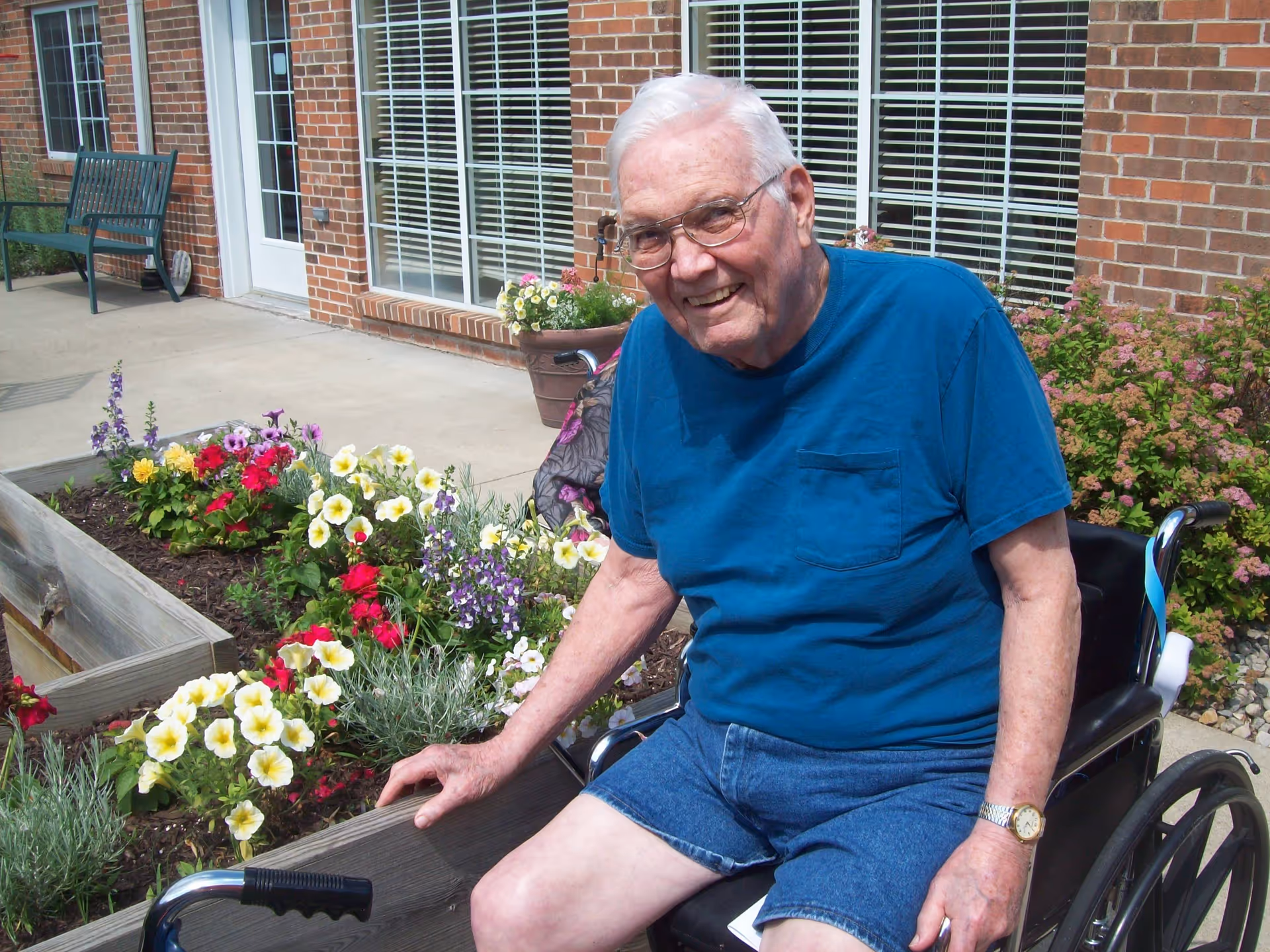 An elderly man with white hair and glasses sitting in a wheelchair outside near a raised flower bed with colorful flowers. He is wearing a blue t-shirt and denim shorts, smiling at the camera. Behind him is a brick building with large windows and a door, along with some outdoor benches and plants.