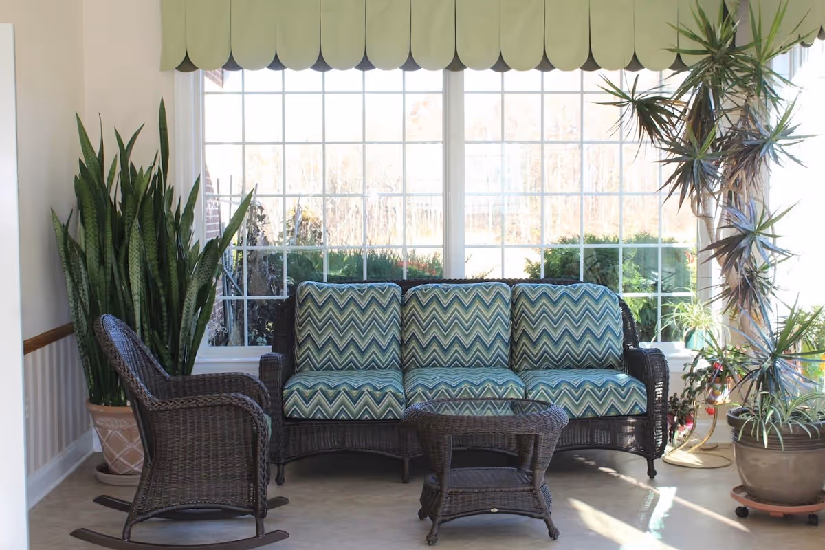 A sunlit seating area with a wicker sofa featuring blue and green chevron-patterned cushions, a matching wicker rocking chair, and a small wicker coffee table. Large windows with a scalloped valance let in natural light, and several potted plants are placed around the seating area.